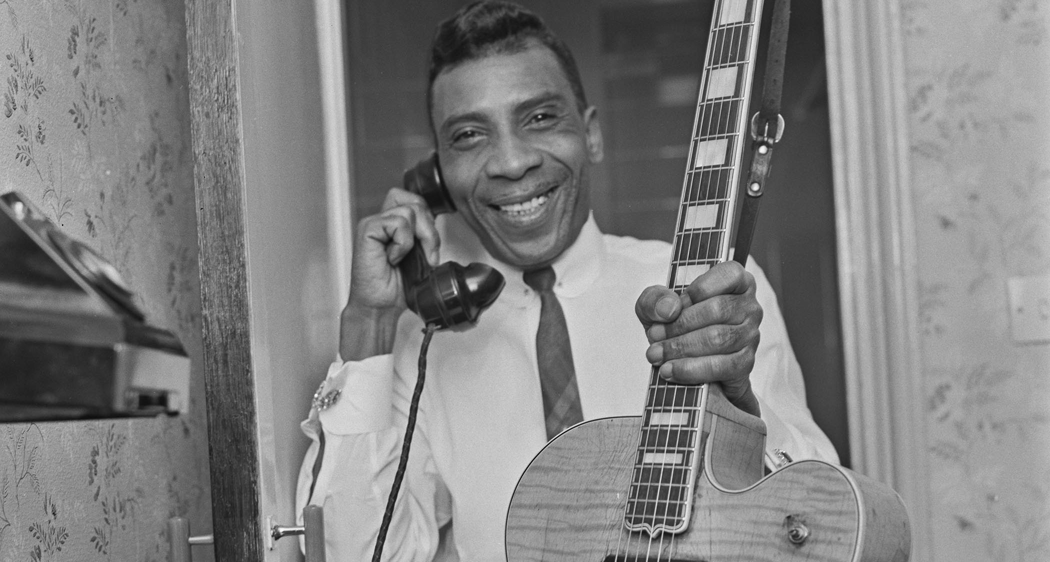 A black-and-white photo of T-Bone Walker answering the phone in one hand and holding his Gibson guitar in another