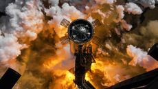 fiery smoke rises from below as a tall chrome cylinder stands next to a tower 