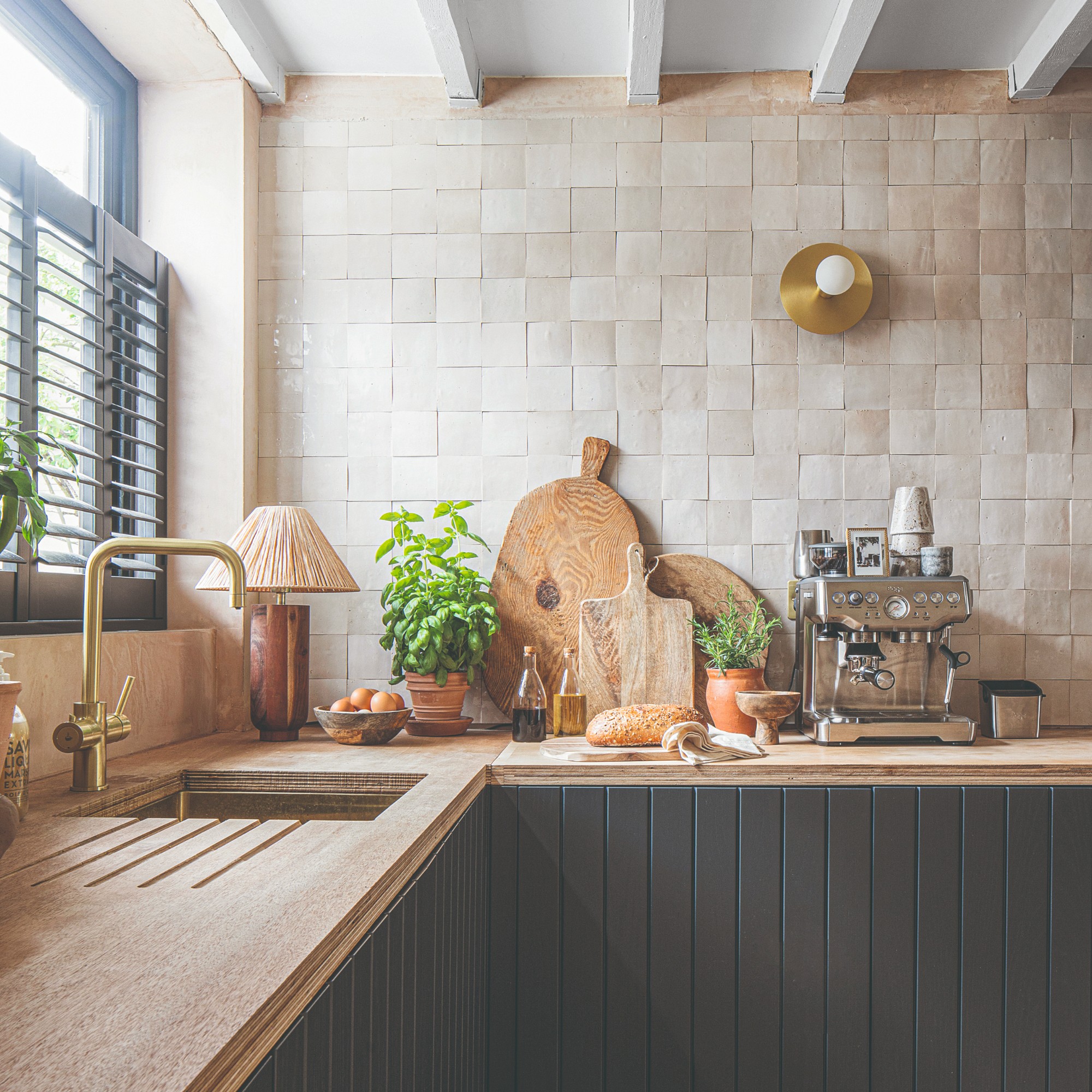 A kitchen with black panelled cabinets, a wooden and rattan wireless table lamp and a round wall sconce on the tiled wall