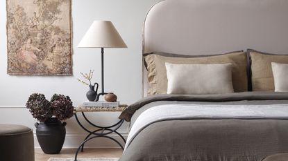 a bedroom with a table lamp, a large bed with neutral bedding and a tapestry on the wall