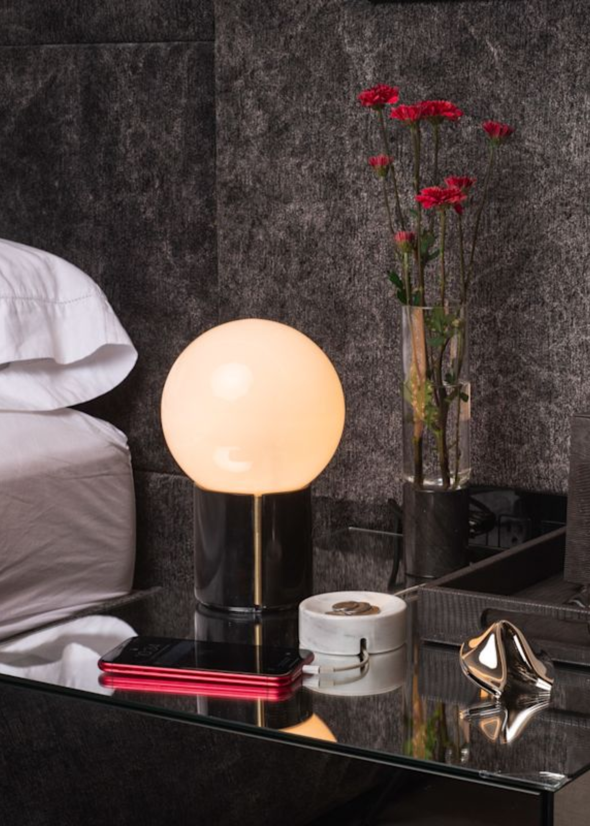 A bedside table with a table lamp, a vase of flowers, a phone, and a gray marble cable holder