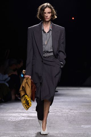 How to wear a suit is shown in a photo of a model on the runway of Bottega Veneta's spring 2025 runway show wearing a gray striped button-down shirt with a dark gray striped blazer, light gray belt, dark gray striped midi skirt, white pumps, a red handbag, and flowers