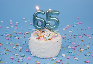 Coconut birthday cake on blue background with burning candle number 65 and many colorful sprinkles.