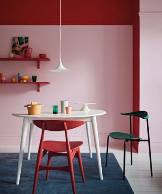 Red and pink painted dining room, red shelving with colorful ornaments and vases, white rounded dining table with red and green dining chairs, low hanging white cone pendant over table, white painted wooden floor, blue carpet