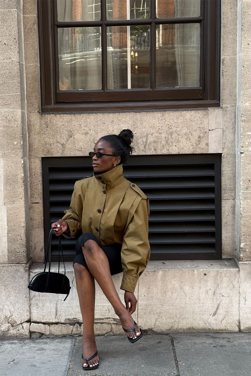 A woman sitting outside on the ledge of a building wearing an olive green funnel neck cropped trench coat styled with black pedal pushers, black heeled thong sandals, a black bowler bag, black oval sunglasses, and silver jewelry