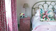 Green, white and pink bedroom with pink curtains, a green bed with pink and white bedding and a botanical print cushion, and a perspex nightstand filled with rose petals with a green lamp and flowers.
