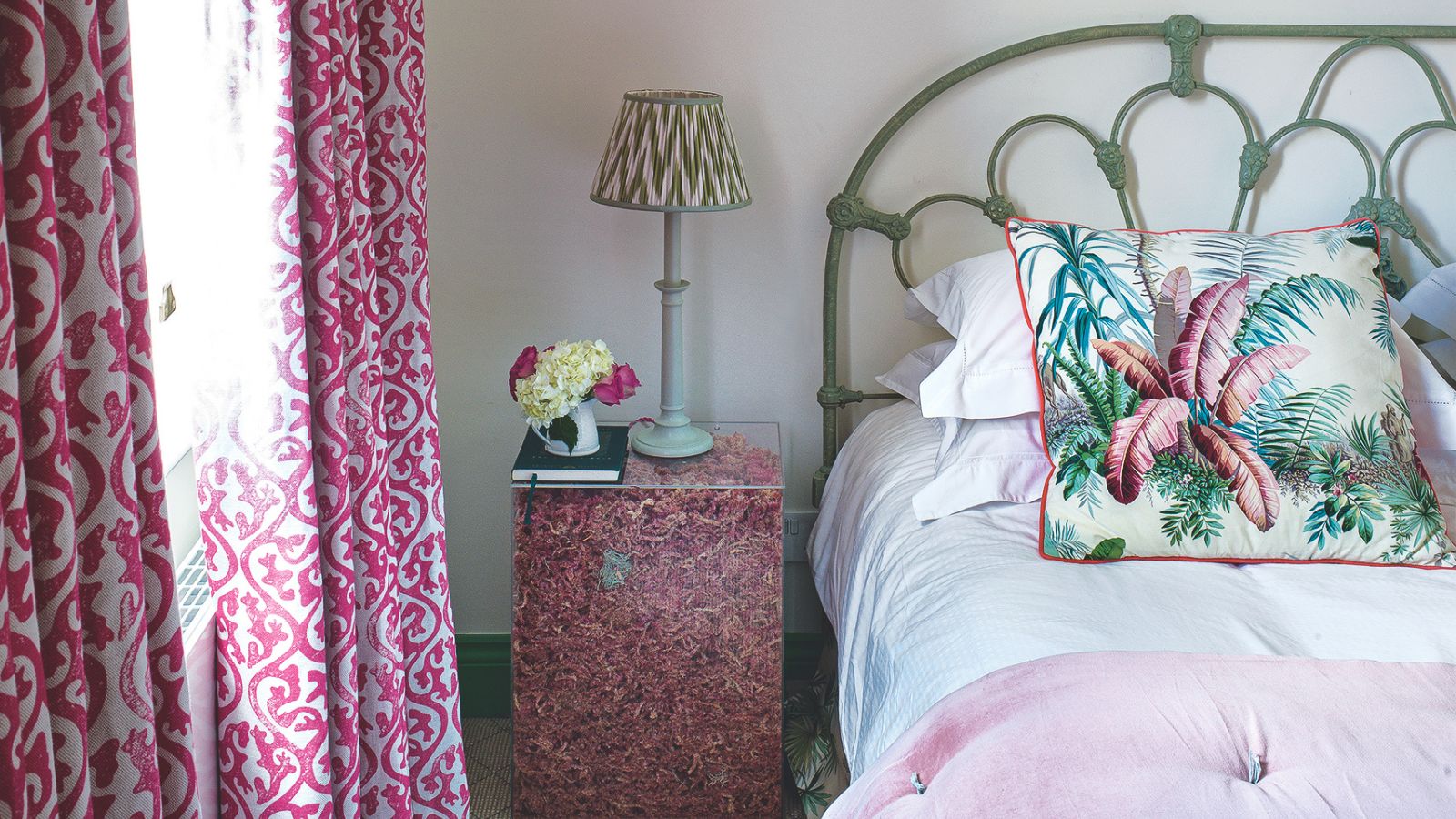 Green, white and pink bedroom with pink curtains, a green bed with pink and white bedding and a botanical print cushion, and a perspex nightstand filled with rose petals with a green lamp and flowers.