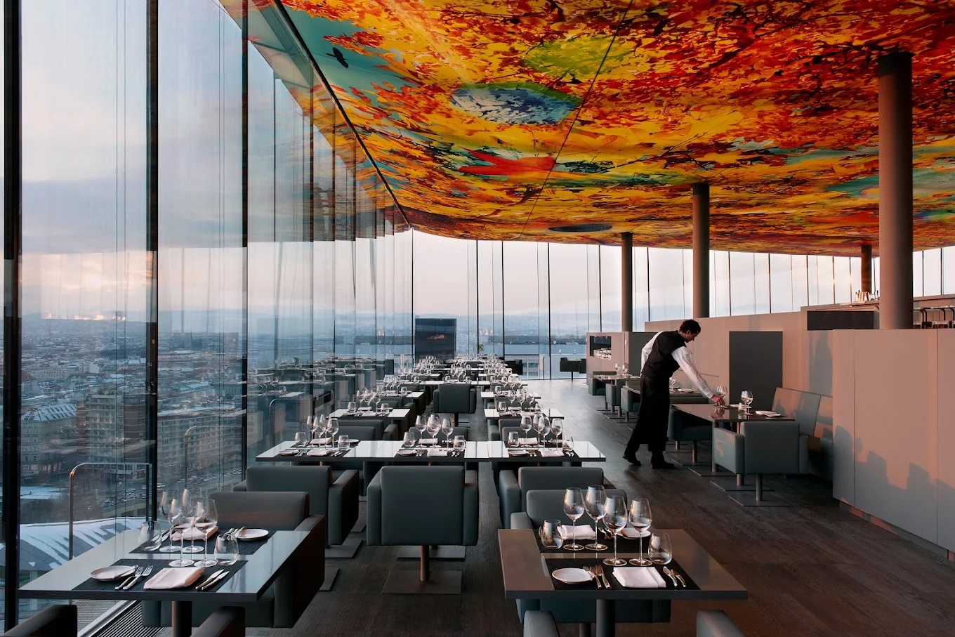A panoramic restaurant rooftop features wrapped-around, floor-to-ceiling windows, a painted ceiling in bright shades, and minimalist tables with gray upholstered seating, all captured at dusk.