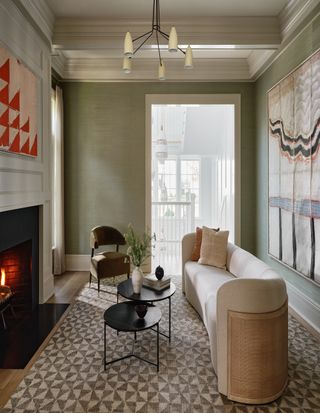 Image of a sage green living room with a white and rattan, curved sofa. There is a black nesting table in front of the sofa. The rug is a geometric, triangular pattern, and there is an artwork hanging over the fireplace that has the same pattern.