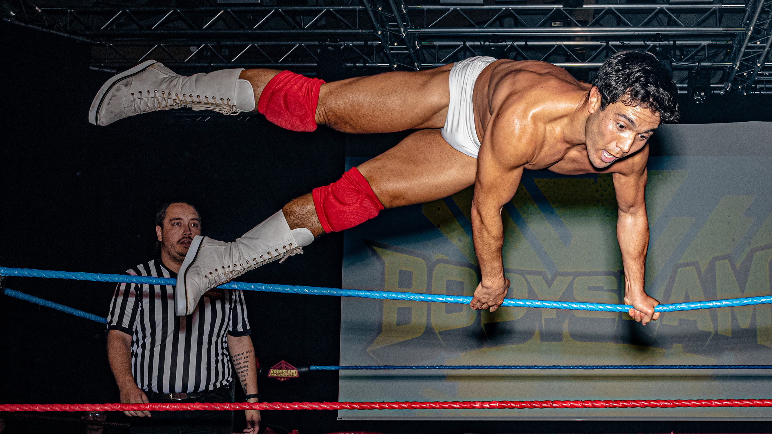 A wrestler in white trunks and red boots leaps mid-air inside a wrestling ring. Another wrestler stands below, surprised. A referee observes the scene