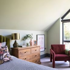 A light green bedroom with a wooden set of drawers and a burgundy armchair next to an olive green bed frame. 