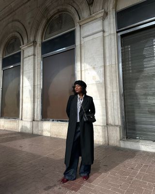 French influencer wears black coat, blue jeans, striped shirt and black hat, standing on Paris street.