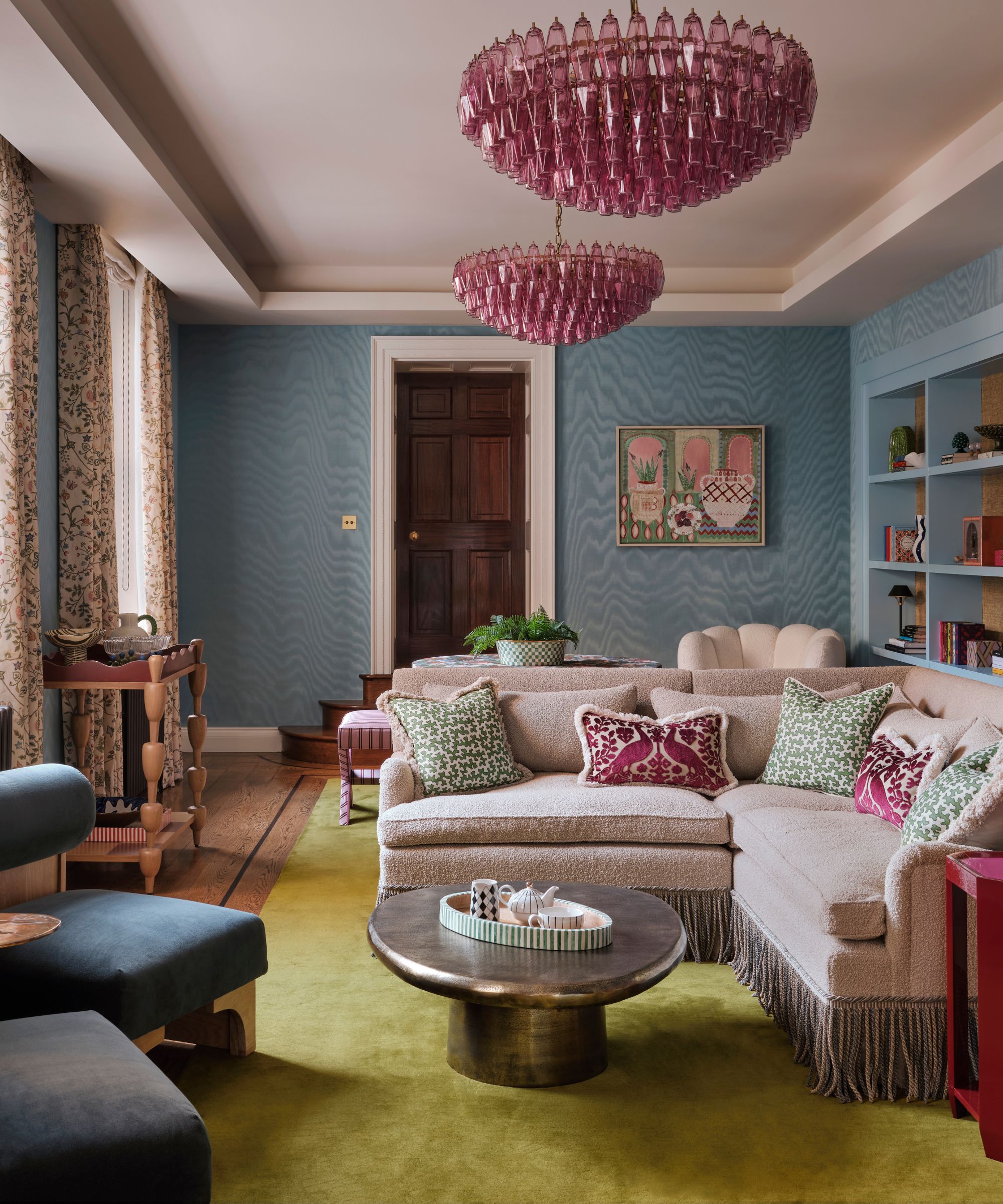 a blure moire wallpapered home library with bookcases on the right, pink chandeliers and a fringed settee sat on a green rug