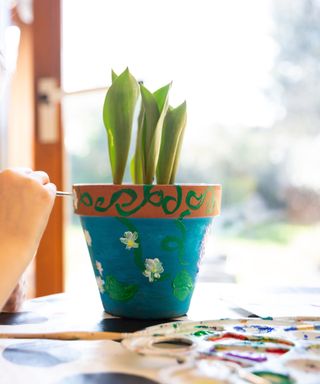 Hand painting white flowers with teal-blue colors in a terracotta flowerpot with three green stems and placing a white paint palette with colorful paints next to the pot