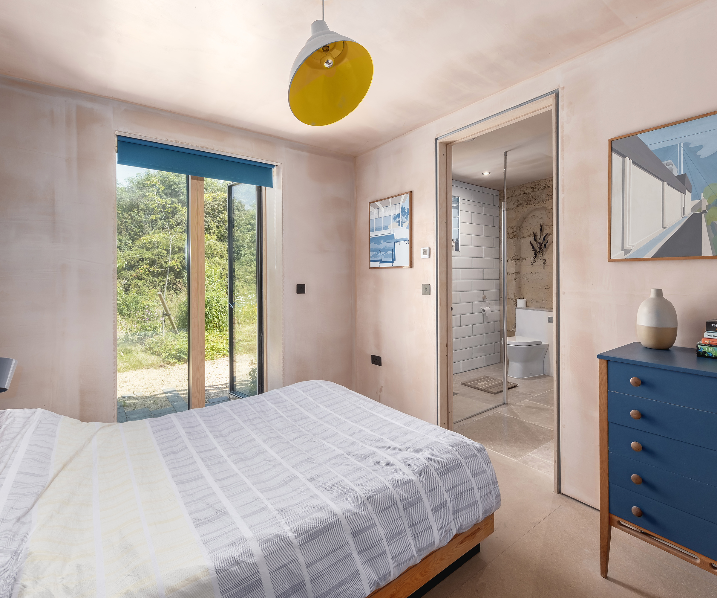 A bedroom in a self build hope with exposed plaster. A door is an open which leads to an ensuite bathroom. There is an open glass door leading out to the garden