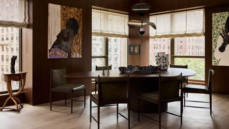 Image of a dark brown wood dining room with windows on the back wall that have sheer linen Roman blinds. There is an organically shaped wooden dining table in the center of the room with black leather dining chairs around it.