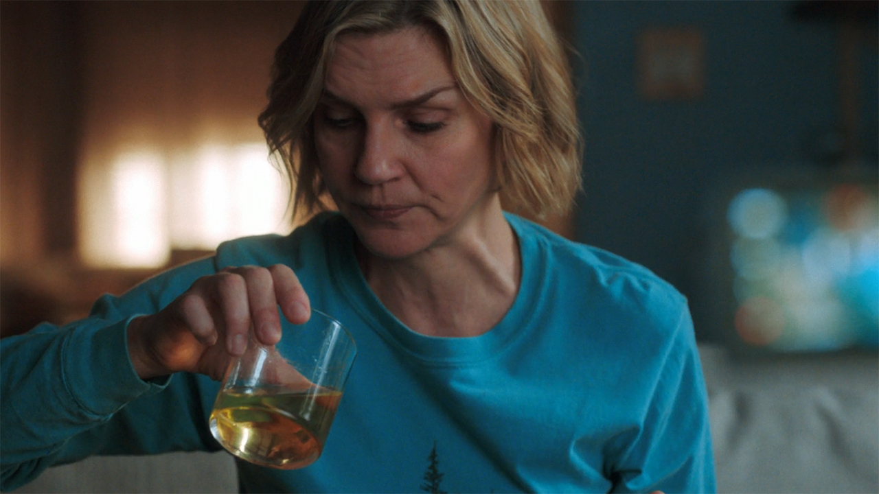 Carol sitting down and looking at an orange liquid in a glass in Pluribus episode 5