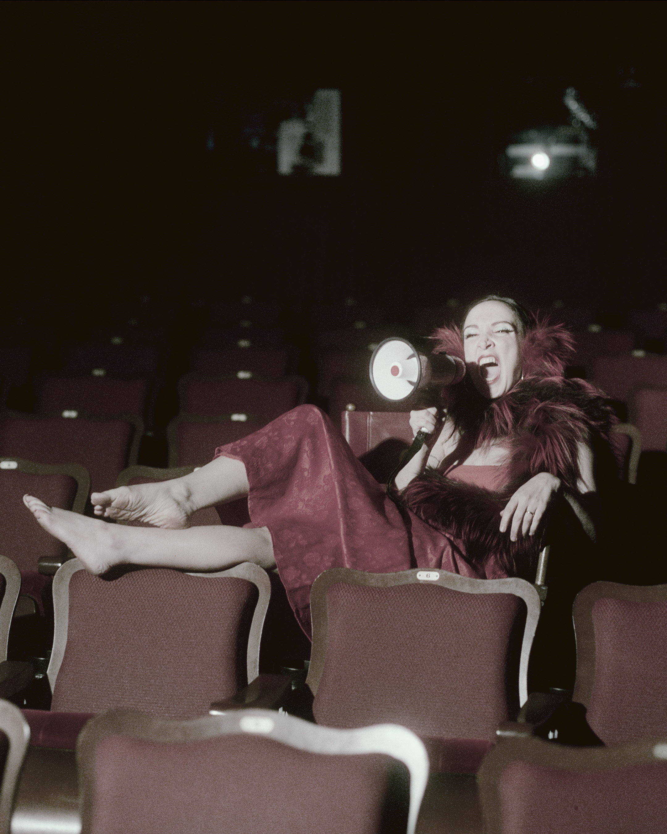 Mary Bronstein lounging barefoot in a theater seat, holding a megaphone and laughing.