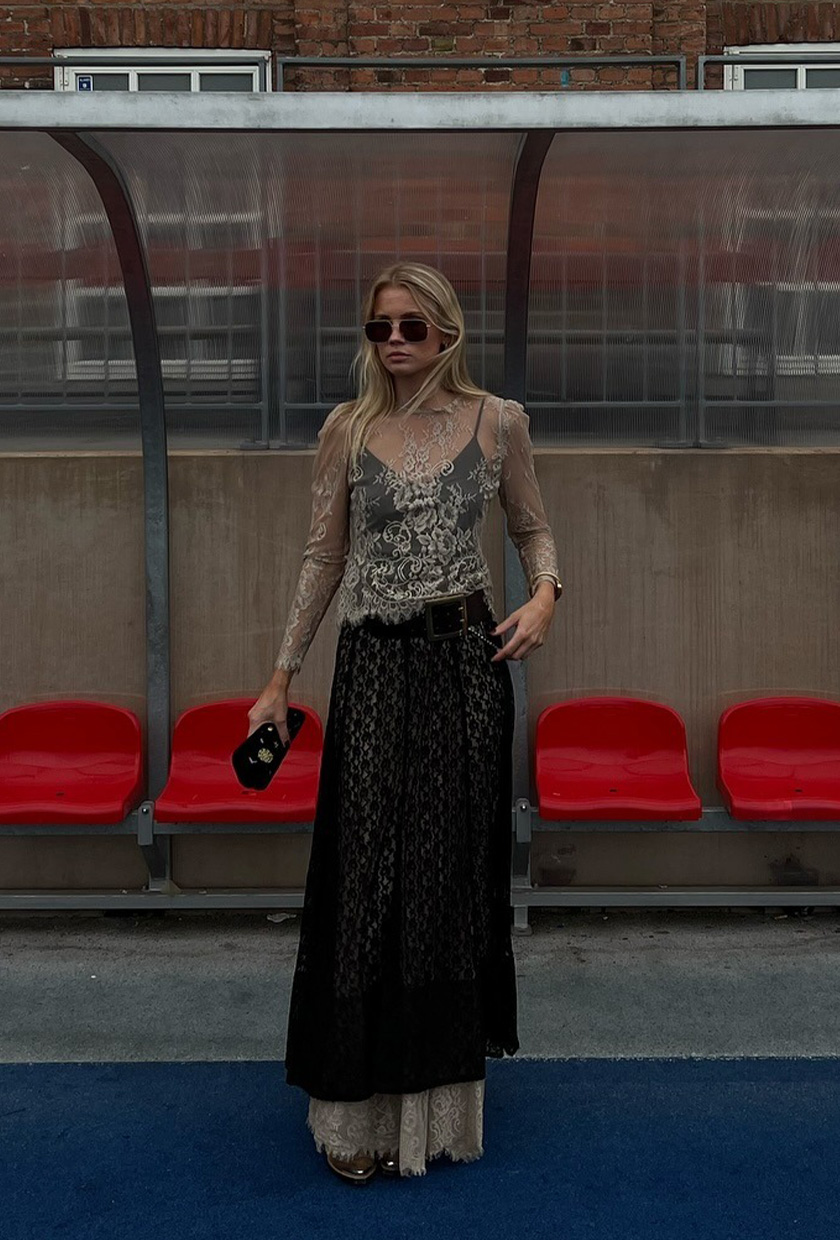 A woman standing outside on a track field wearing a light tan sheer lace shirt layered over a black sheer lace dress layered over a tan sheer lace skirt, gold flats, a black belt, gold bangles, aviator sunglasses, and a black clutch bag.