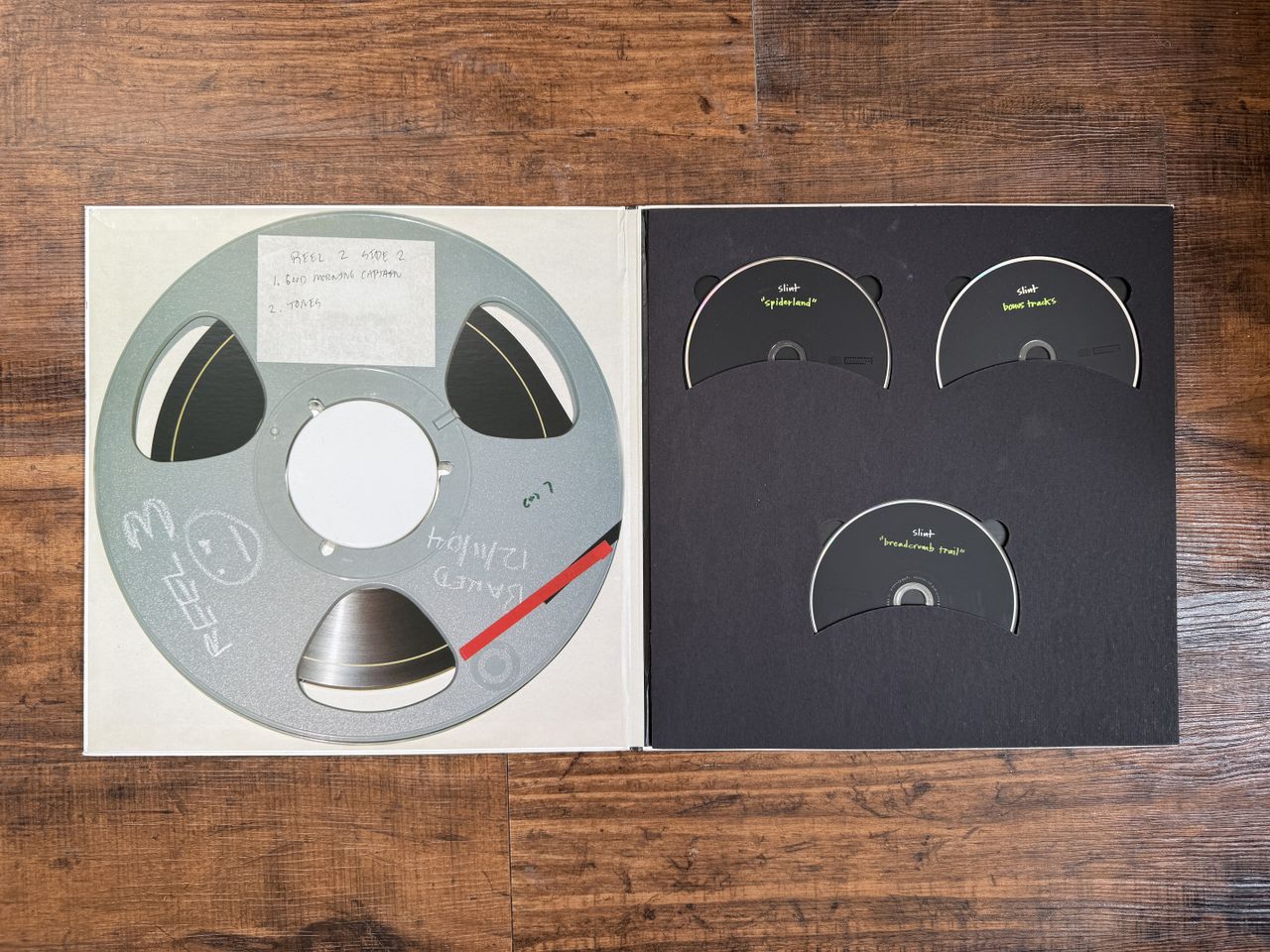 The inside of a gatefold vinyl copy of Spiderland by Slint. The left side shows a tape reel, while the right houses three discs. It is placed on a wooden floor.