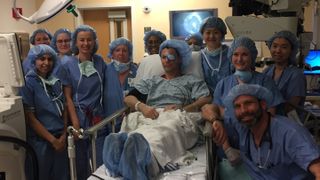 A photo of a patient with their surgical team after surgery. The patient is sat on a hospital bed and the team is gathered around him. 