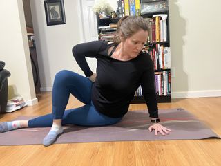 Jennifer Rizzuto performs a seated spinal mobility move on a yoga mat in a living room. She is dressed in a sports top and leggings and sits on the floor. Her left leg lies straight on the floor and her right leg crossed over it, while her torso is propped upright by her left arm. Her right arm is bent and her right hand rests on her hip.