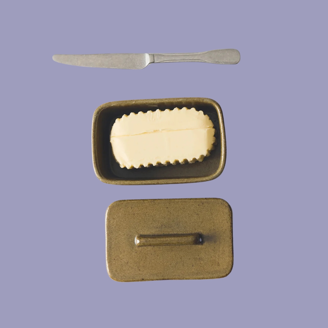 A pale purple background is topped by a ceramic, moss green butter dish filled with butter, opened up, and with a silver knife next to it.