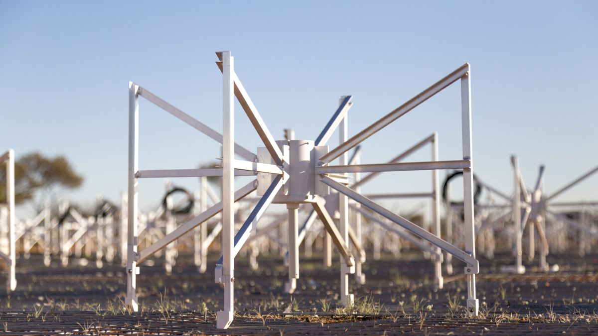 A white contraption stands on the ground surrounded by many others. It has four straight legs attached to a central cylinder by three support struts each.