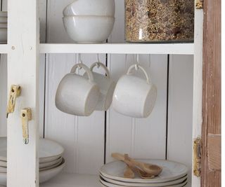 White ceramic mugs hanging from hooks under a cabinet shelf with nested white plates and wooden spoons in view