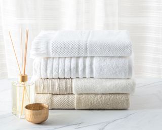 Pile of beige and white towels next to a reed diffuser and small copper bowl on a white marble counter with white candle behind