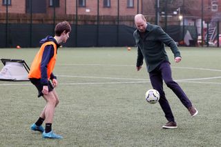 Prince William playing soccer in sneakers and a green shacket with a teenage boy on a grass pitch