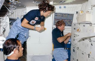 three astronauts in blue t shirts work on board nasa's space shuttle