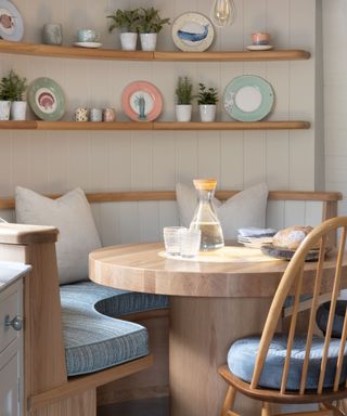 Cosy breakfast nook with built-in wooden banquette seating, blue upholstered cushions, a round oak table, and open shelves displaying decorative plates and plants