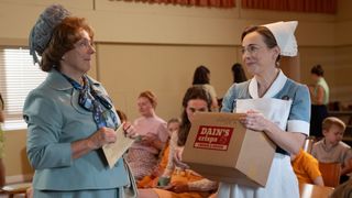 Nurse Shelagh holding a cardboard box and smiling at Violet, who is looking smart in a pale blue suit and hat