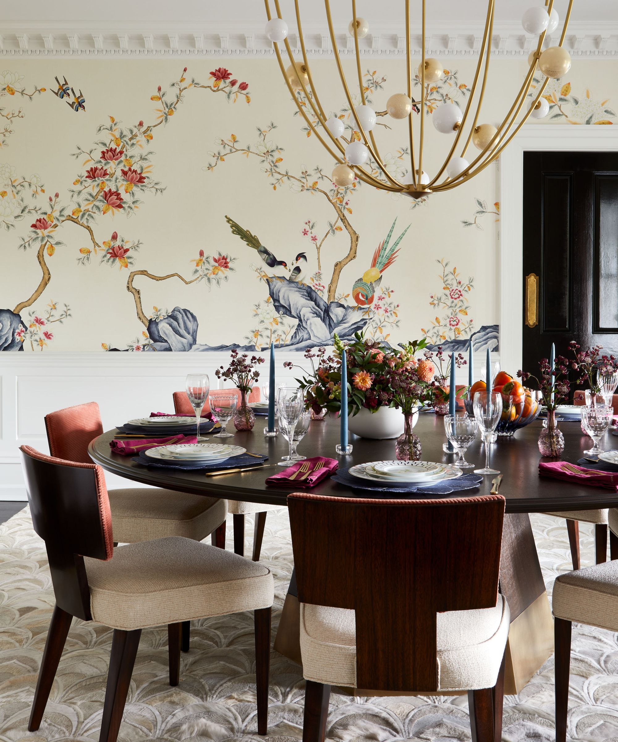 Dining room with ornate wallpaper and half-paneled walls. A large circular wooden table sits in the middle with wooden chairs upholstered in cream. A large gold chandelier hangs above