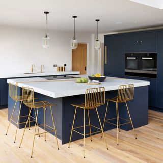 blue kitchen island with white worktops and gold stools, blue cabinets and white walls