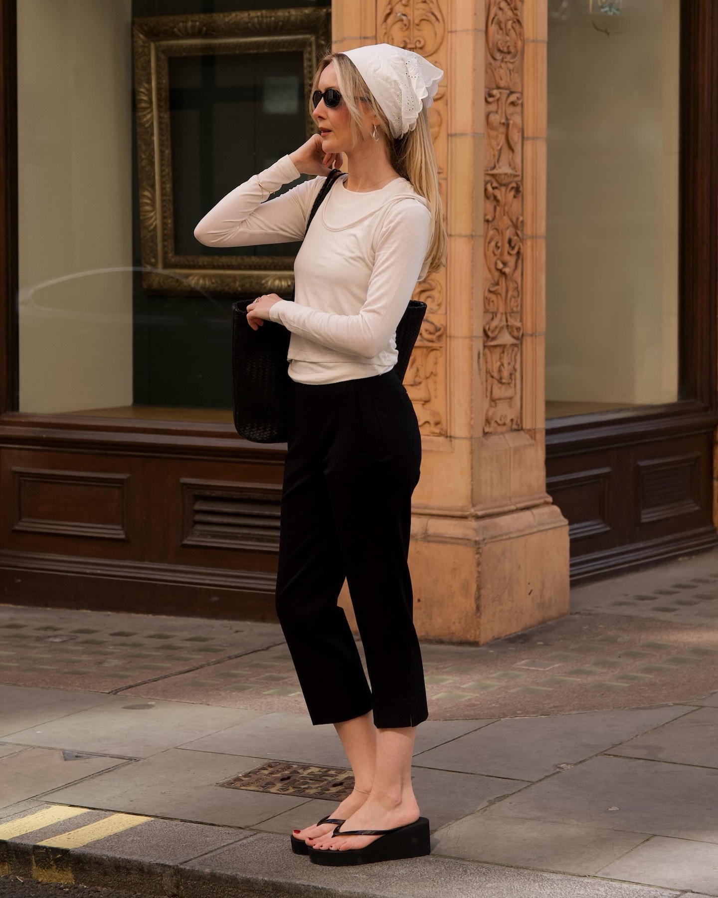 British style influencer Chloe Kath Butler poses on a sidewalk wearing a white eyelet head scarf, sunglasses, a layered ivory long-sleeve tee, black tote bag, capri pants, and wedge flip-flop sandals