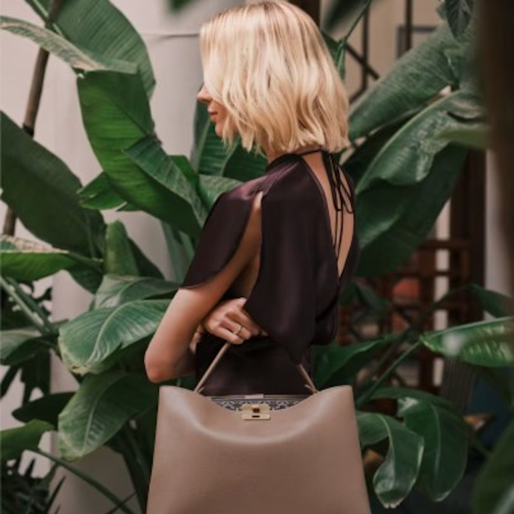 A profile photo of a woman modelling a black satin tie-back top while crossing her arms holding a beige-pink handbag in front of a large indoor plant