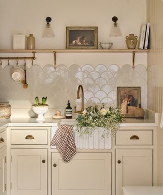 cream painted kitchen with cream units, wooden shelf, scalloped edge cream tiles, glass wall lights and flowers in sink