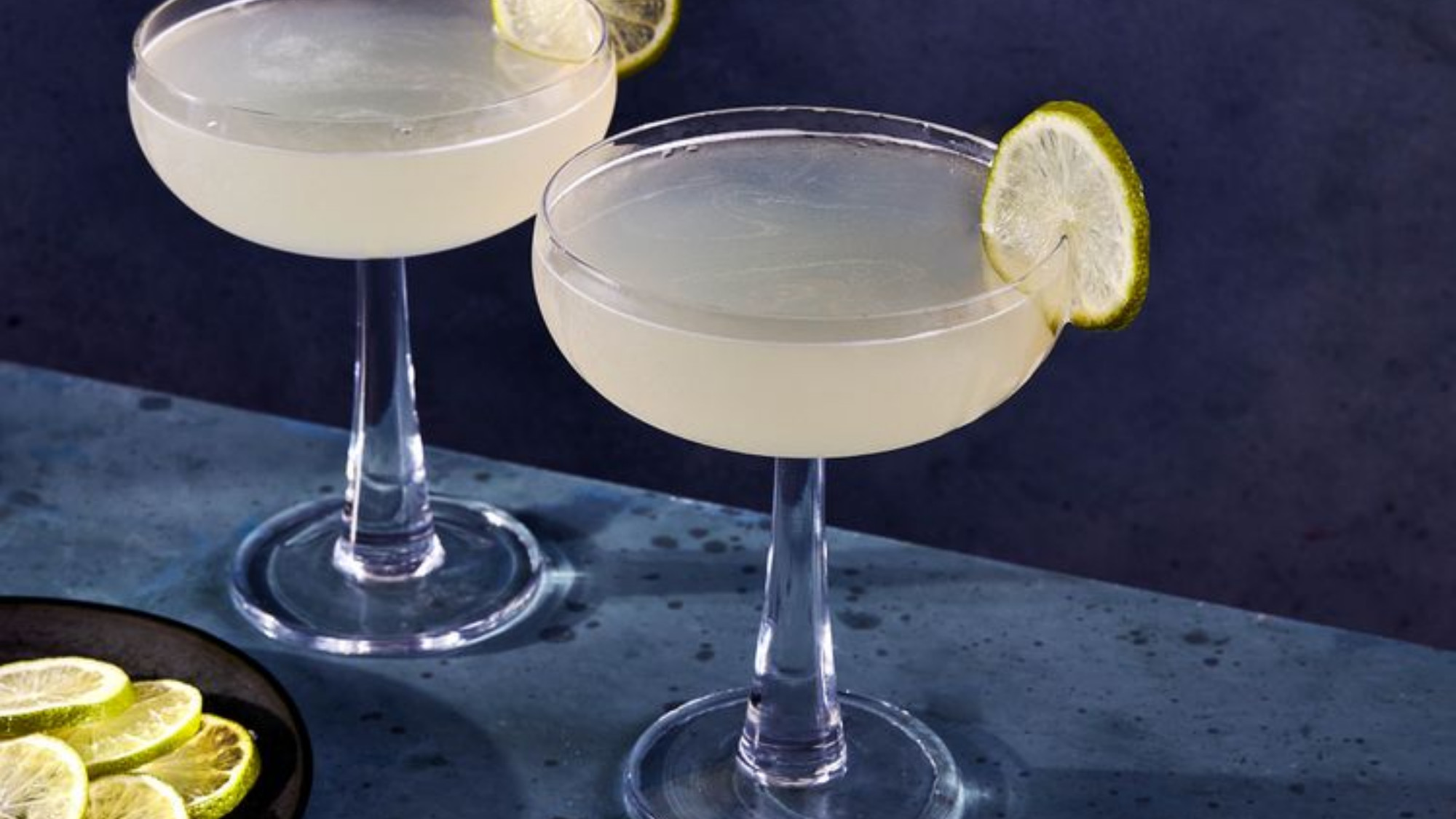 side shot of two martini glasses filled with gimlets against a blue surface and blue wall