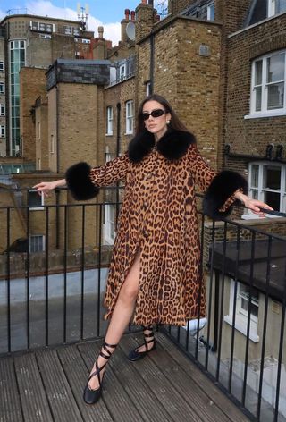 The leopard print coat trend is shown on a woman standing outside wearing black sunglasses, gold earrings, a leopard print coat, and lace-up ballet flats