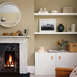 Living room with a lit fire in a traditional fireplace, shelves and a cupboard, a large leather armchair and cosy painted walls