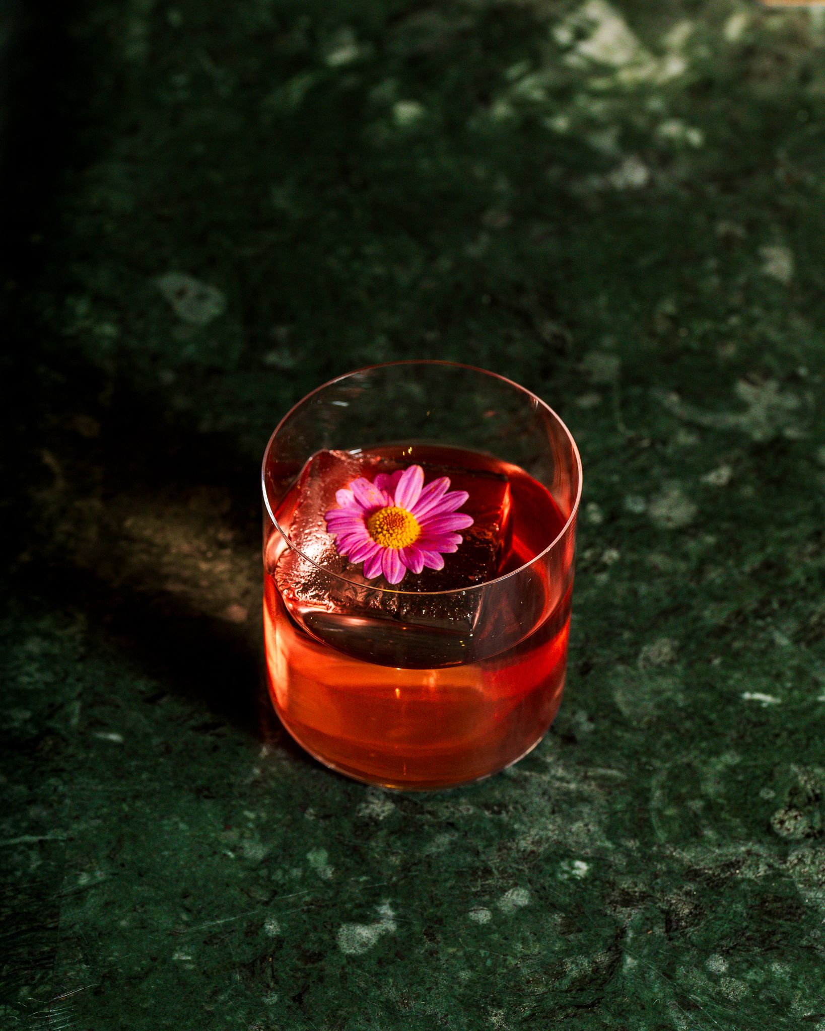 Two red cocktails served in transparent glasses sitting on terrazzo green, or blue, tables, respectively.