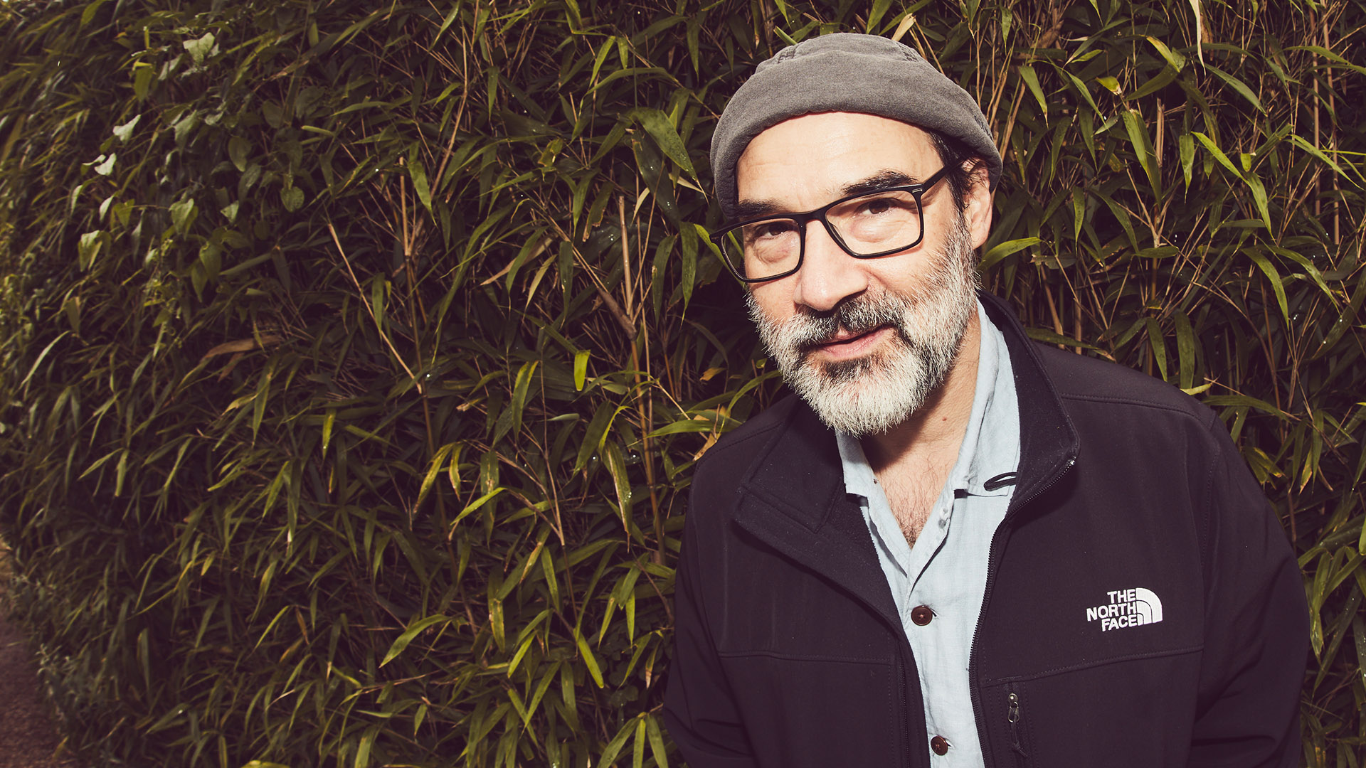 A photo of Adam Buxton in front of some leafy bushes. 
