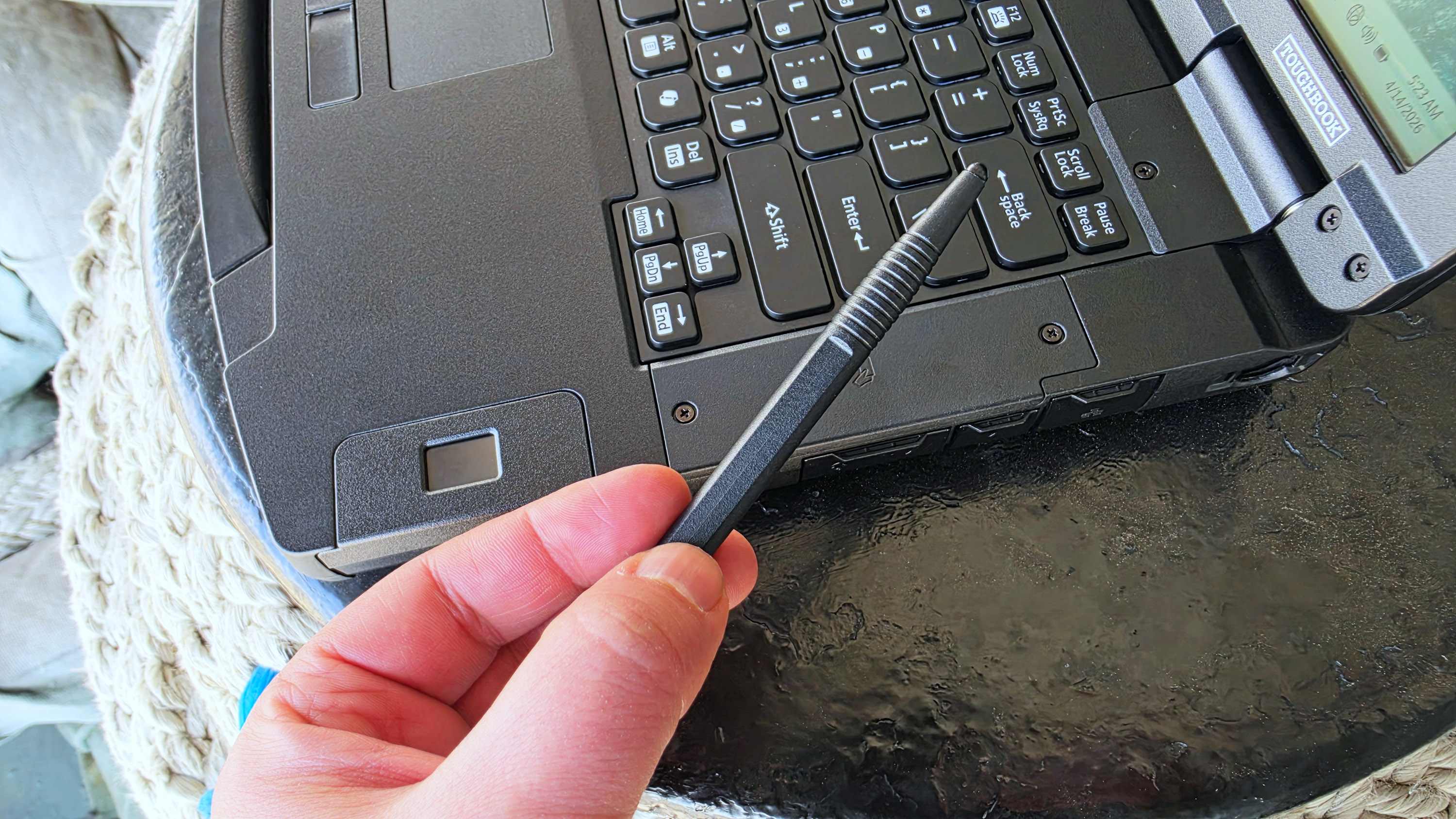 A hand holds a black stylus above the keyboard of a rugged laptop on a round, textured table, suggesting a setting for tech work or repair.