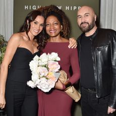 Meghan Markle wearing a black corset and pants with her arm around Kadi Lee who is holding a bouquet of roses, and Serge Normant standing next to them in a black jacket