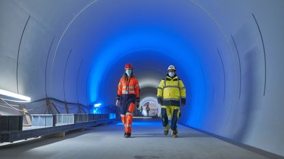 A woman and a man wearing hardhats and construction suits walk down a tunnel lit with blue light