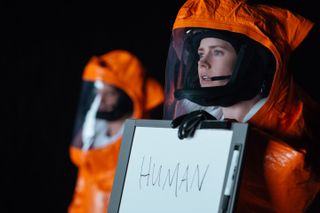 A woman in an orange hazmat suit holds up a whiteboard with the word "human" written on it.