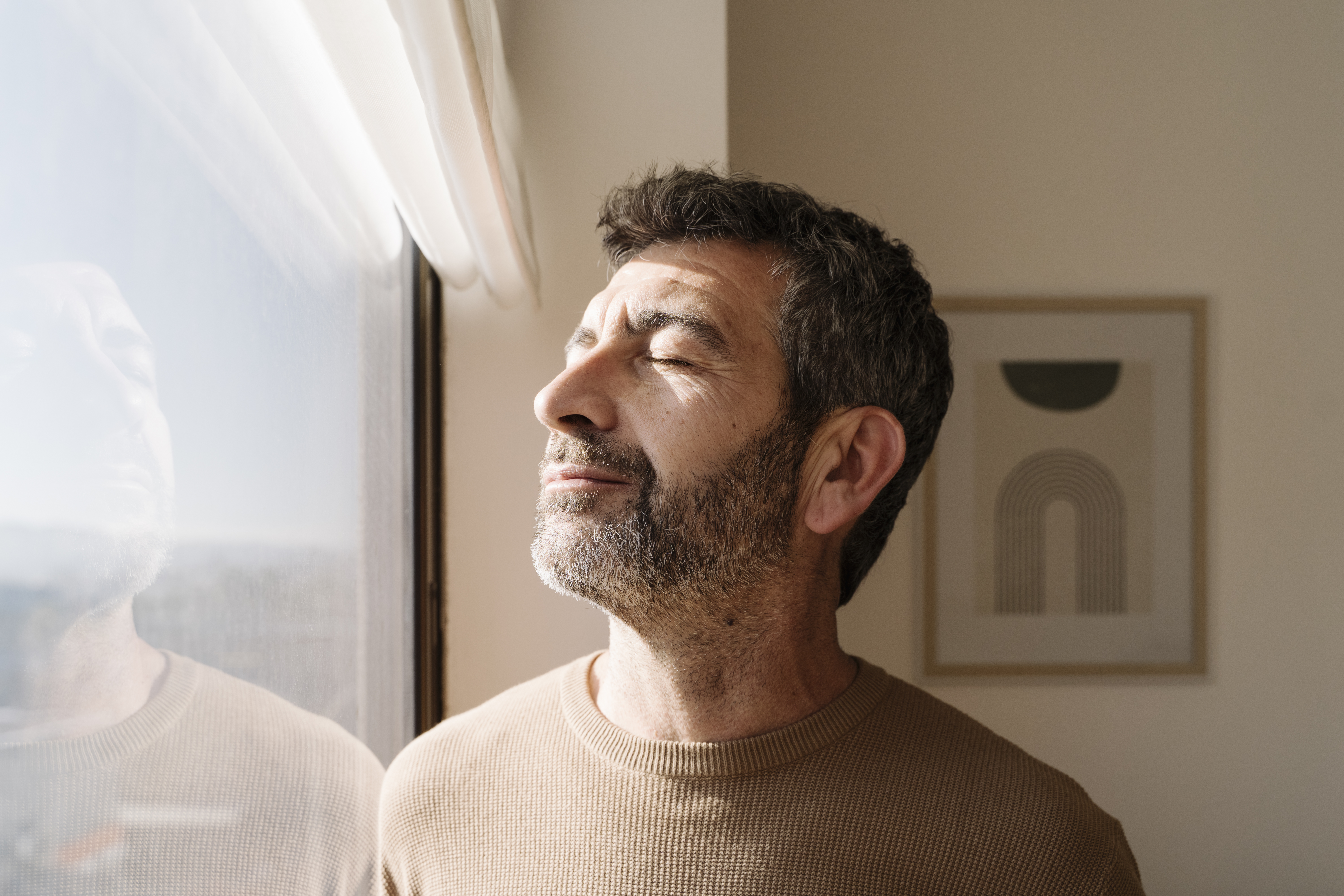 A man standing beside a closed window indoors on a sunny day.