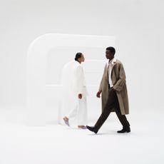 Two models passing eachother next to a bit "Ff" statue with white studio background one wearing a long trench coat and the other wearing an all white trouser suit sold at Farfetch